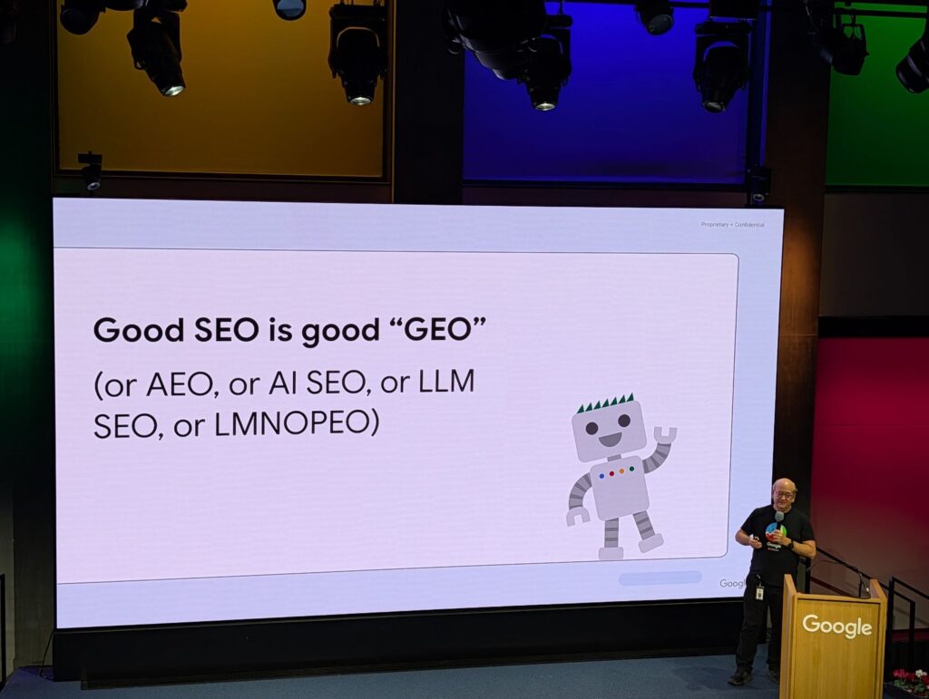 Foto einer Präsentationsfolie auf einer Google-Bühne. Auf der Folie steht «Good SEO is good “GEO” (or AEO, or AI SEO, or LLM SEO, or LMNOPEO)». Rechts ist eine einfache Roboterillustration zu sehen. Vor der Leinwand steht John Mueller von Google am Rednerpult und präsentiert den Inhalt.